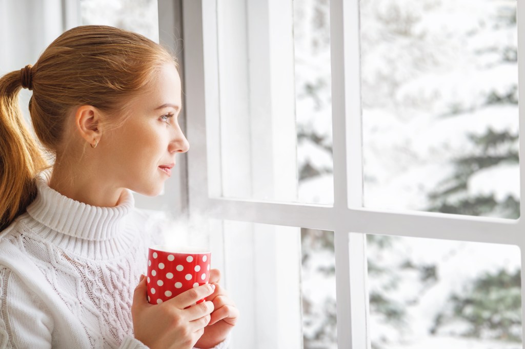 A person wrapped in a warm blanket holding a hot mug of tea by a sunny window with snow outside, illustrating ways to beat winter blues and save on heating bills.